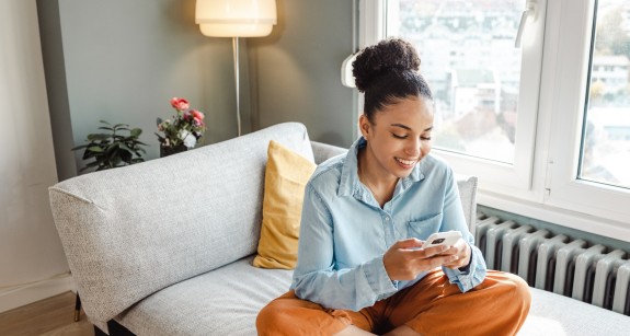 Junge Frau sitzt lächelnd auf einem Sofa und schaut in ihr Handy