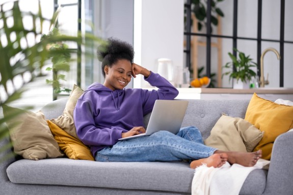junge Frau mit lila Pulli sitzt mit Laptop auf der Couch