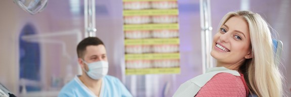 junge blonde Frau sitzt auf dem Zahnarztstuhl, Zahnarzt im Hintergrund